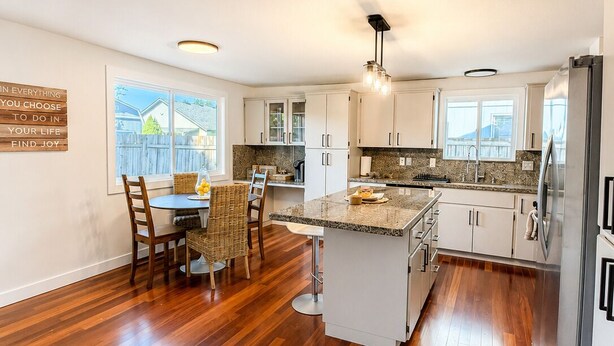 A modern kitchen with wooden flooring, white cabinets, a central island, and a dining area with wicker chairs. - 4 BD, Pet Friendly - Modern chic beautiful, fenced yard (Vancouver)