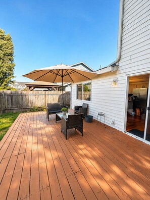 Eine Holzterrasse mit Gartenmöbeln, einem großen Sonnenschirm und einer Glasglastür, die zu einer Küche führt.