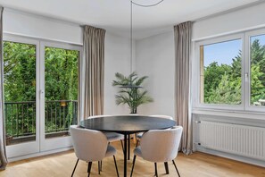 En matplats med ett runt bord, fyra stolar, en krukväxt och stora fönster med utsikt över grönområden.