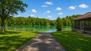 Blick auf einen See mit einem Schotterweg, der zu einer Holzterrasse mit Seeblick führt, umgeben von üppiger Vegetation und blauem Himmel.