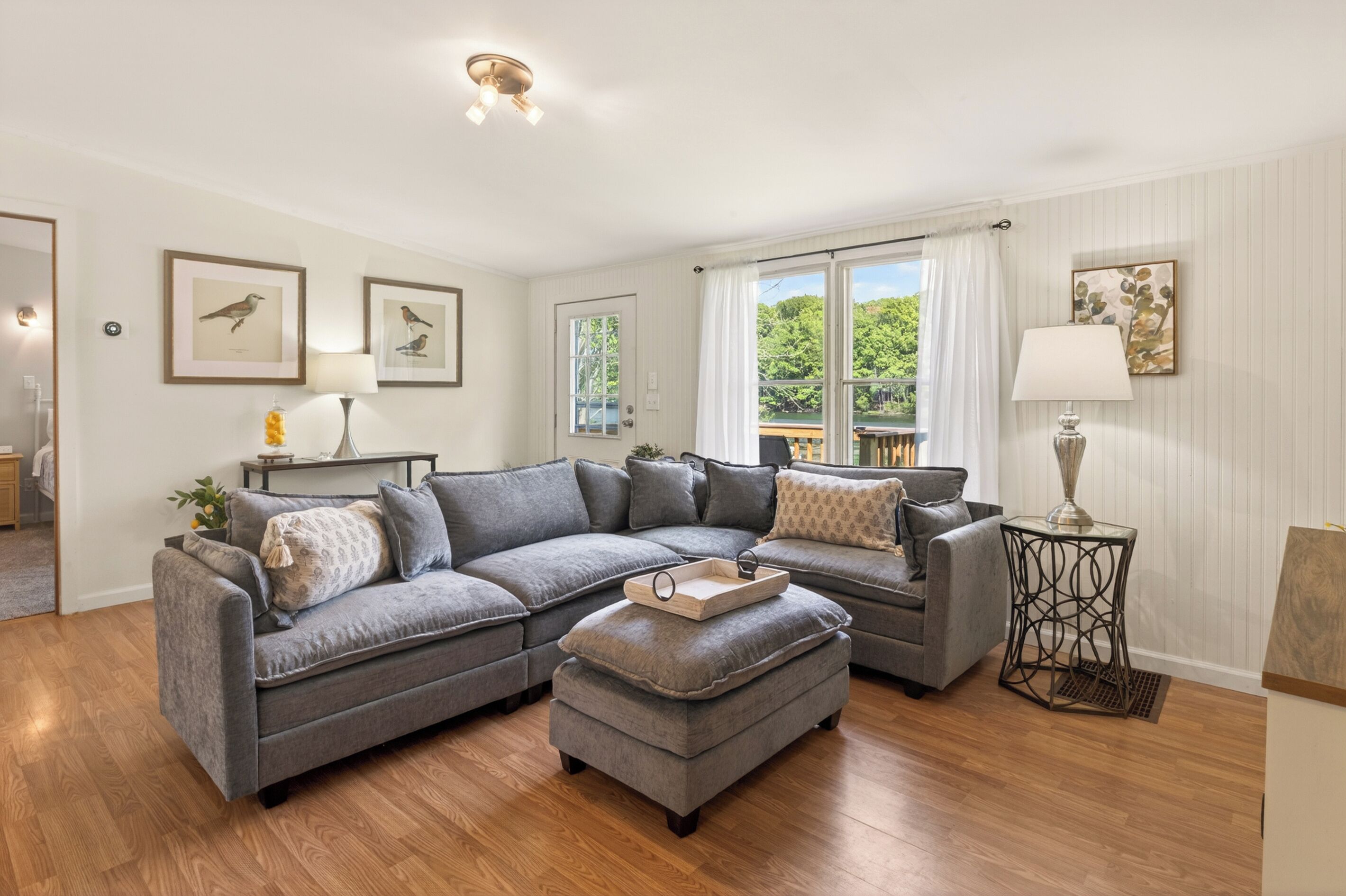 A living room with a grey sectional sofa, a small ottoman, a side table, framed wall art, and a window with curtains.