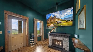 Ein Zimmer mit Kamin aus Stein, Holzdielenboden, einer Tür mit Ausblick und einem großen Landschaftsbild an der Wand.