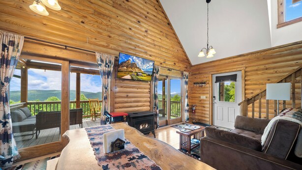 Ein Holzblockhaus-Interieur mit Holzbalkendecke, Kamin, Sofa, Tisch und Blick durch große Fenster.