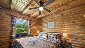 Ein Schlafzimmer in einer Holzhütte mit Bett, Deckenventilator, Fenster mit Vorhängen und Nachttisch mit Lampe.