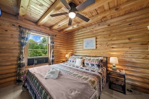 Ein Schlafzimmer in einer Holzhütte mit Bett, Deckenventilator, Fenster mit Vorhängen und Nachttisch mit Lampe.