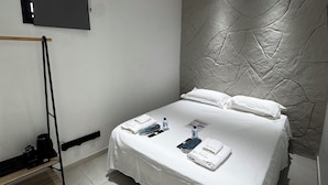A modern hotel room with a bed, a flat-screen TV mounted on the wall, a towel rack, and a small shelf. - 8-enclosure room - BS HOUSE HÔTEL (Pointe-à-Pitre)