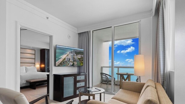 A hotel room with a bed, a TV, a desk, a chair, a sofa, and a balcony with a view of the ocean. - Sorrento 1514 (Miami Beach)