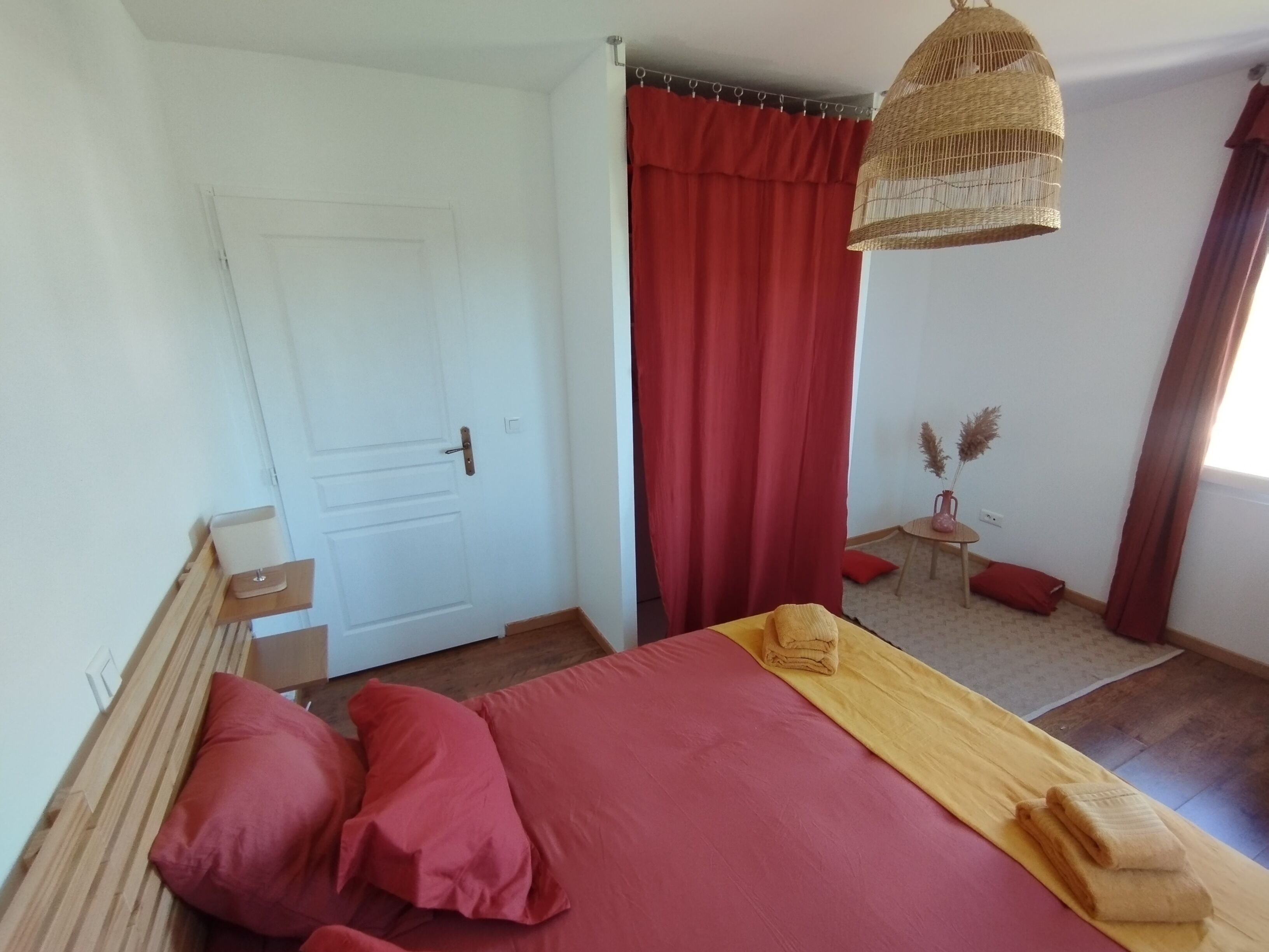 A bedroom with a bed, red curtains, a wooden headboard, a small table with a vase, and a hanging light fixture.