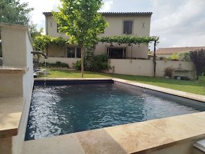 A swimming pool with a tiled edge, a tree, and a building with a tiled roof in the background. - Country house facing the Ventoux (Modène)