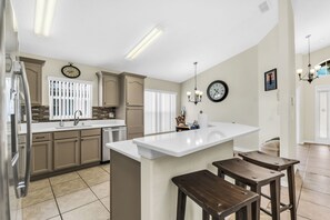 A kitchen with a white island, wooden stools, beige cabinets, a stainless steel refrigerator, and a clock on the wall. - Disney World Retreat Oasis, Pool, Privacy & Comfort. (Kissimmee)