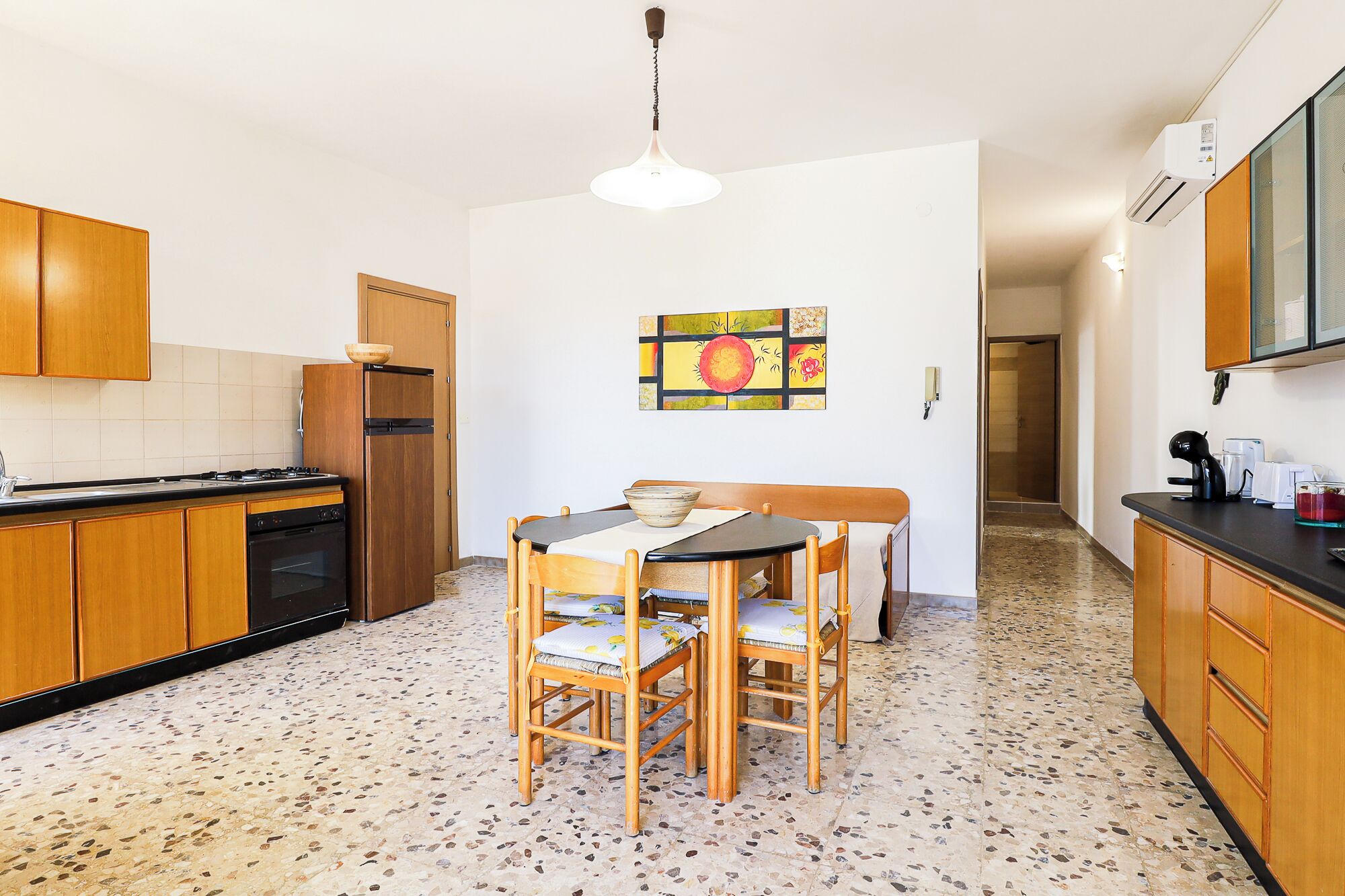 Une cuisine avec des meubles de rangement en bois, une table à manger avec des chaises, un réfrigérateur et un luminaire suspendu.