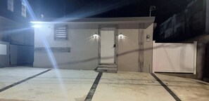 Entrada de un edificio con puerta, dos luces en la pared, unos escalones y una zona vallada.
