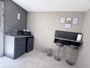 Cocina compacta con lavamanos, microondas y cafetera. Hay dos taburetes de bar y un cubo de basura.