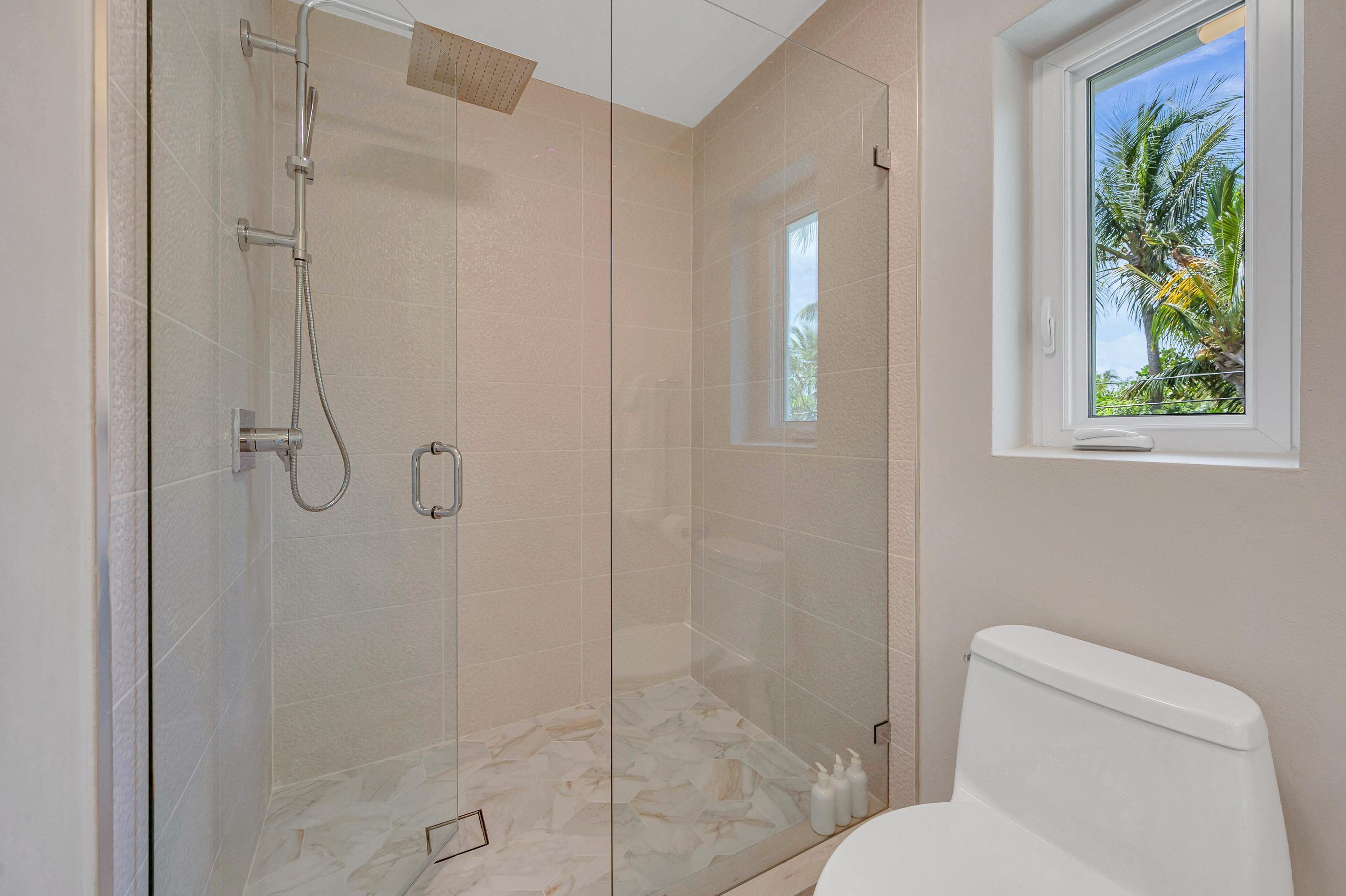 Een moderne badkamer met een douchecabine van glas, een toilet en een raam met uitzicht op palmbomen.