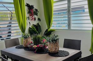 A dining area with a table set for four, surrounded by wicker chairs, with potted plants and a view of the outside.