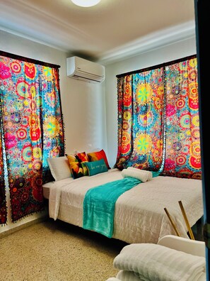 Un dormitorio con cama, cortinas coloridas, un cojín con texto y un equipo de aire acondicionado.