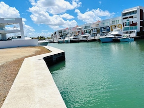 A marina with boats docked, a concrete walkway, and colorful buildings in the background.