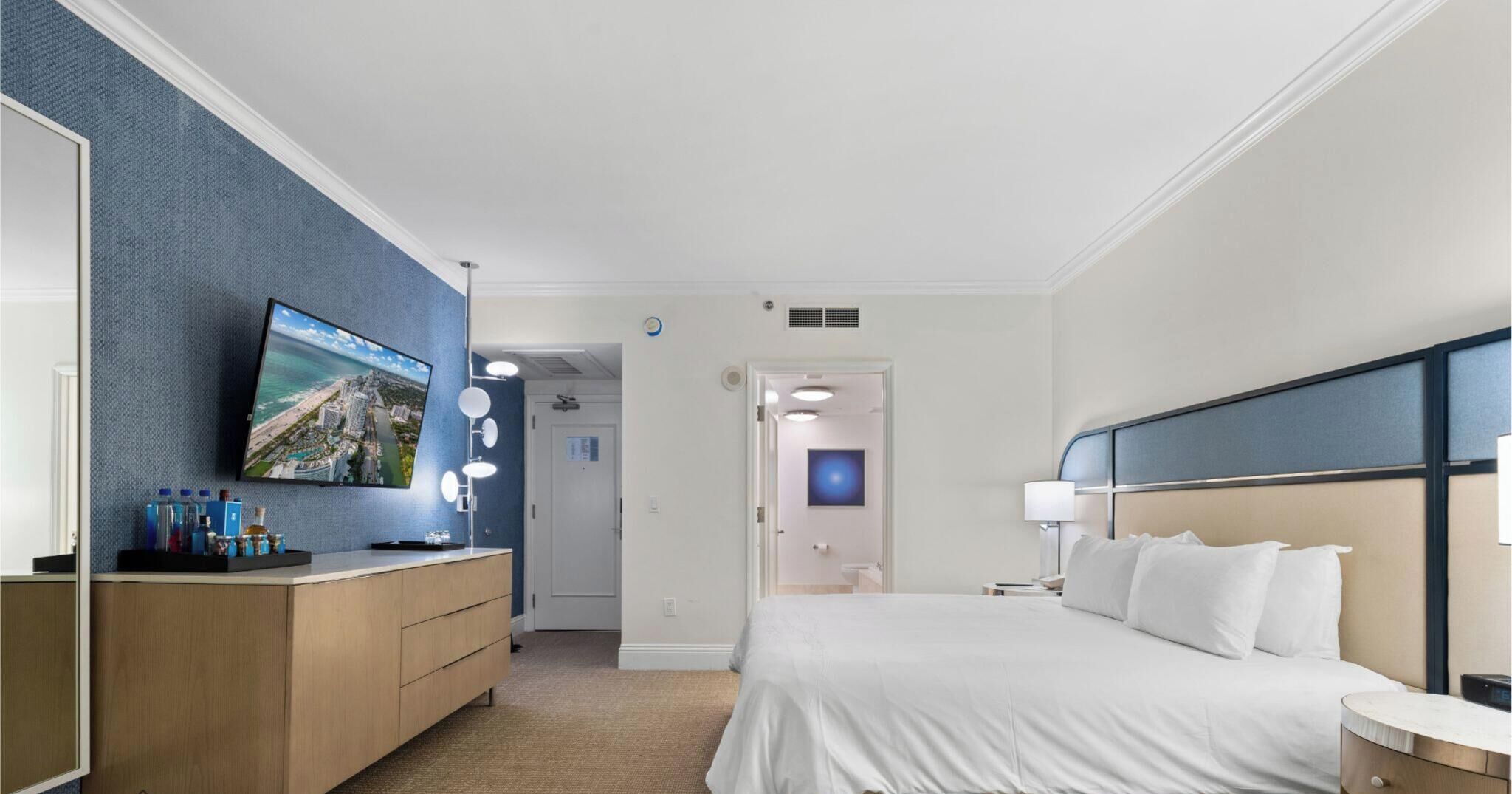 A hotel room with a bed, a TV mounted on a blue wall, a vanity with a mirror, and a door leading to another room.