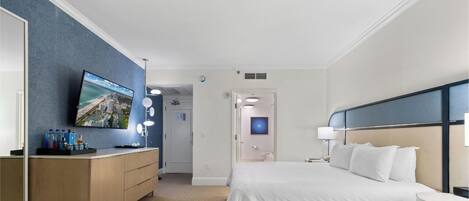 A hotel room with a bed, a TV mounted on a blue wall, a vanity with a mirror, and a door leading to another room.