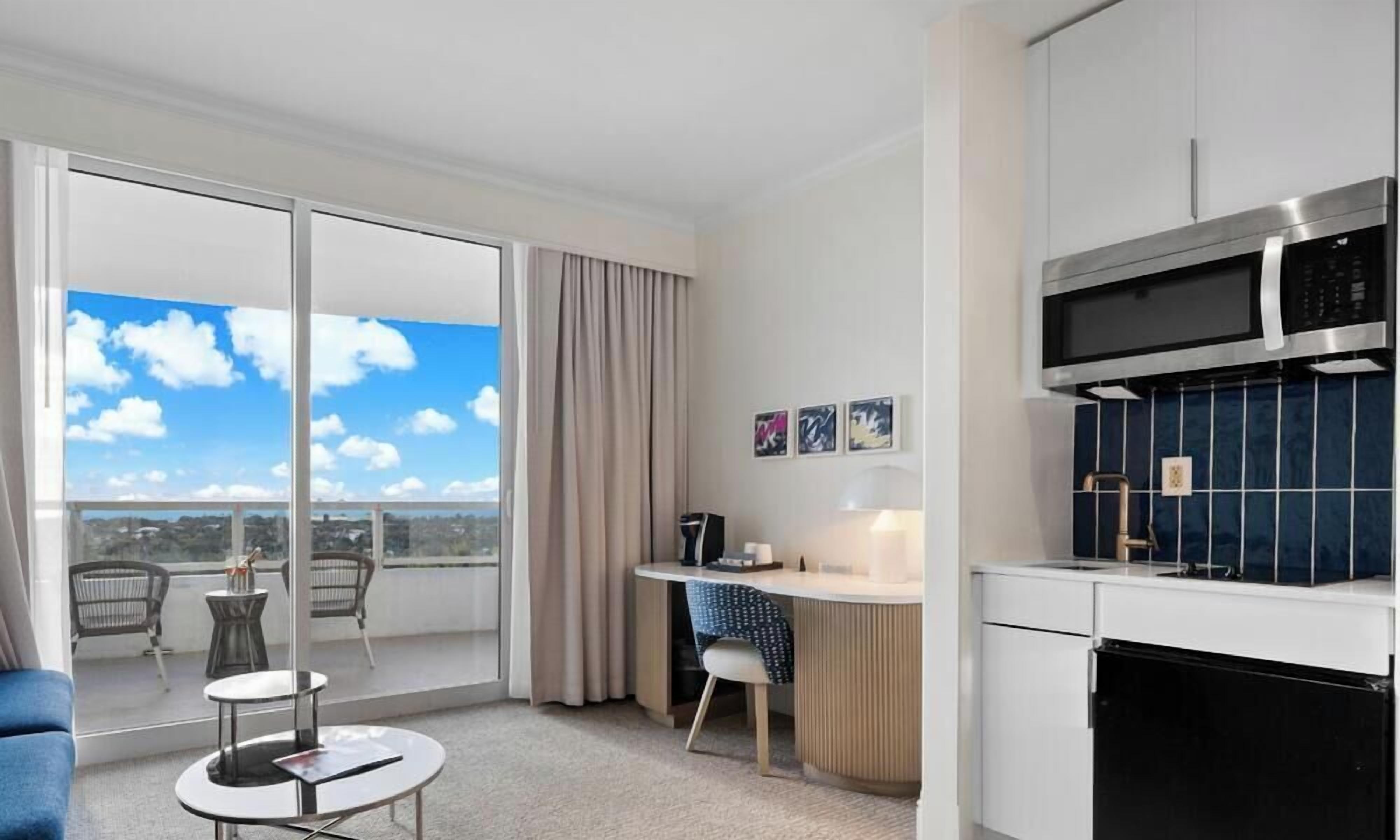 A hotel room with a balcony, a kitchenette, a desk with a chair, and a sofa.