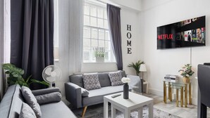 Una sala de estar moderna con un sofá gris, una mesa de centro blanca, un televisor montado en la pared y un ventanal con cortinas.