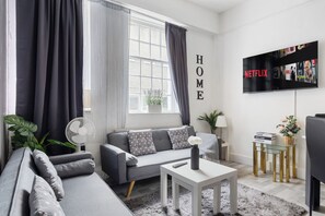 Un soggiorno moderno con un divano grigio, un tavolino da caffè bianco, una TV a parete e una finestra con tende.