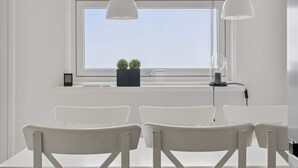 Una habitación con un gran ventanal, dos lámparas colgantes blancas, una pequeña planta en maceta y una mesa blanca con cuatro sillas.