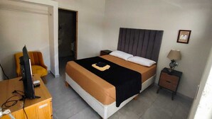 A hotel room with a bed, a desk with a computer, a chair, a bedside table with a lamp, and a door.