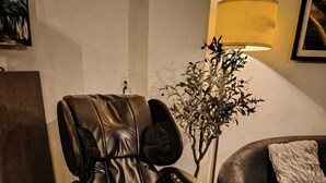 A massage chair in a room with a tiled floor, a lamp, and a couch with a cushion.