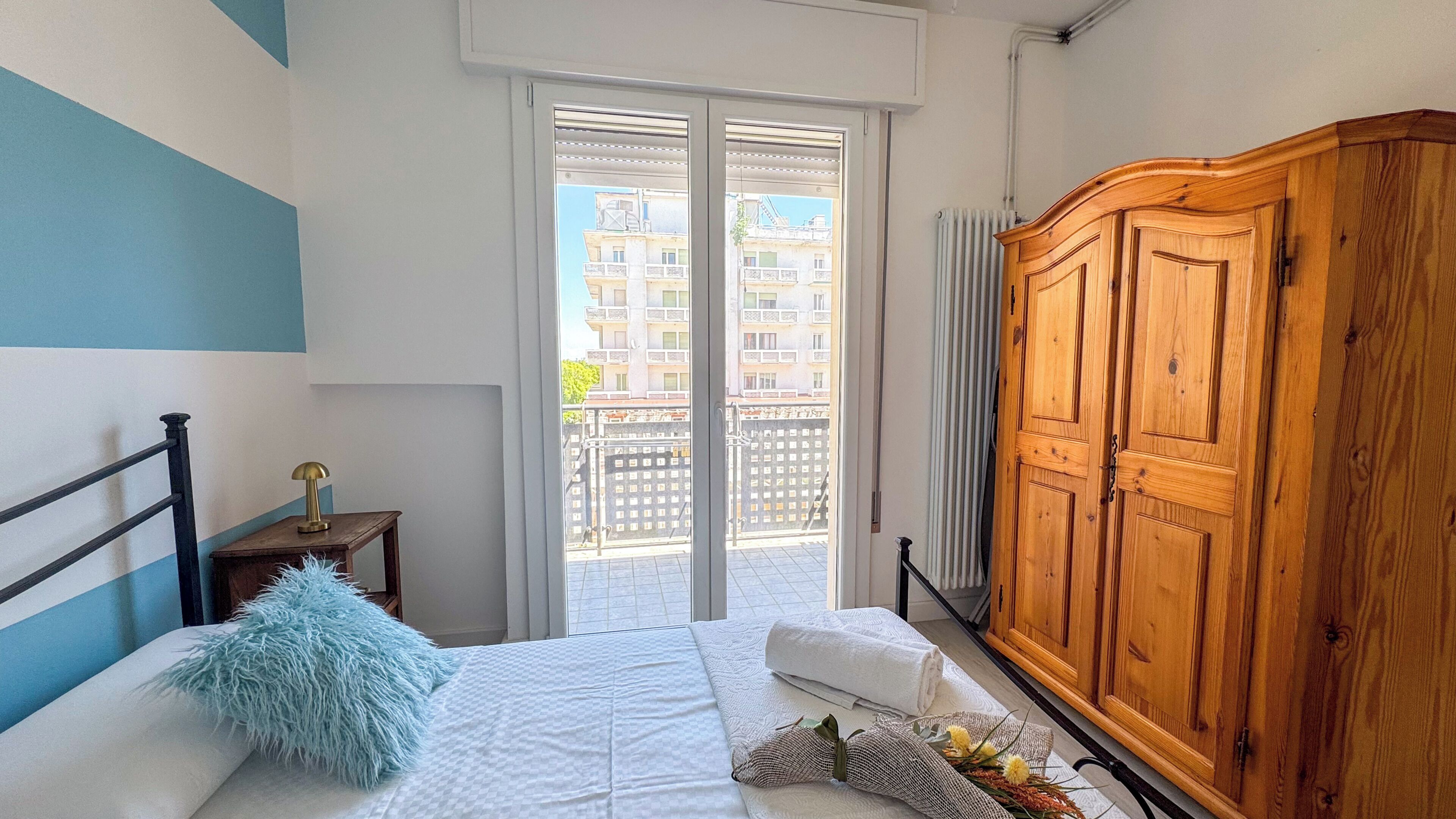 A bedroom with a bed, a wooden wardrobe, a nightstand, a fluffy pillow, and a view of buildings through the window.