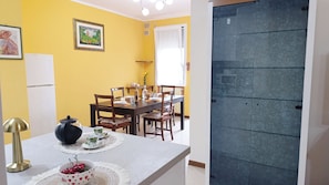 Zona pranzo con tavolo apparecchiato per quattro, sedie, frigorifero bianco e un quadro appeso al muro.