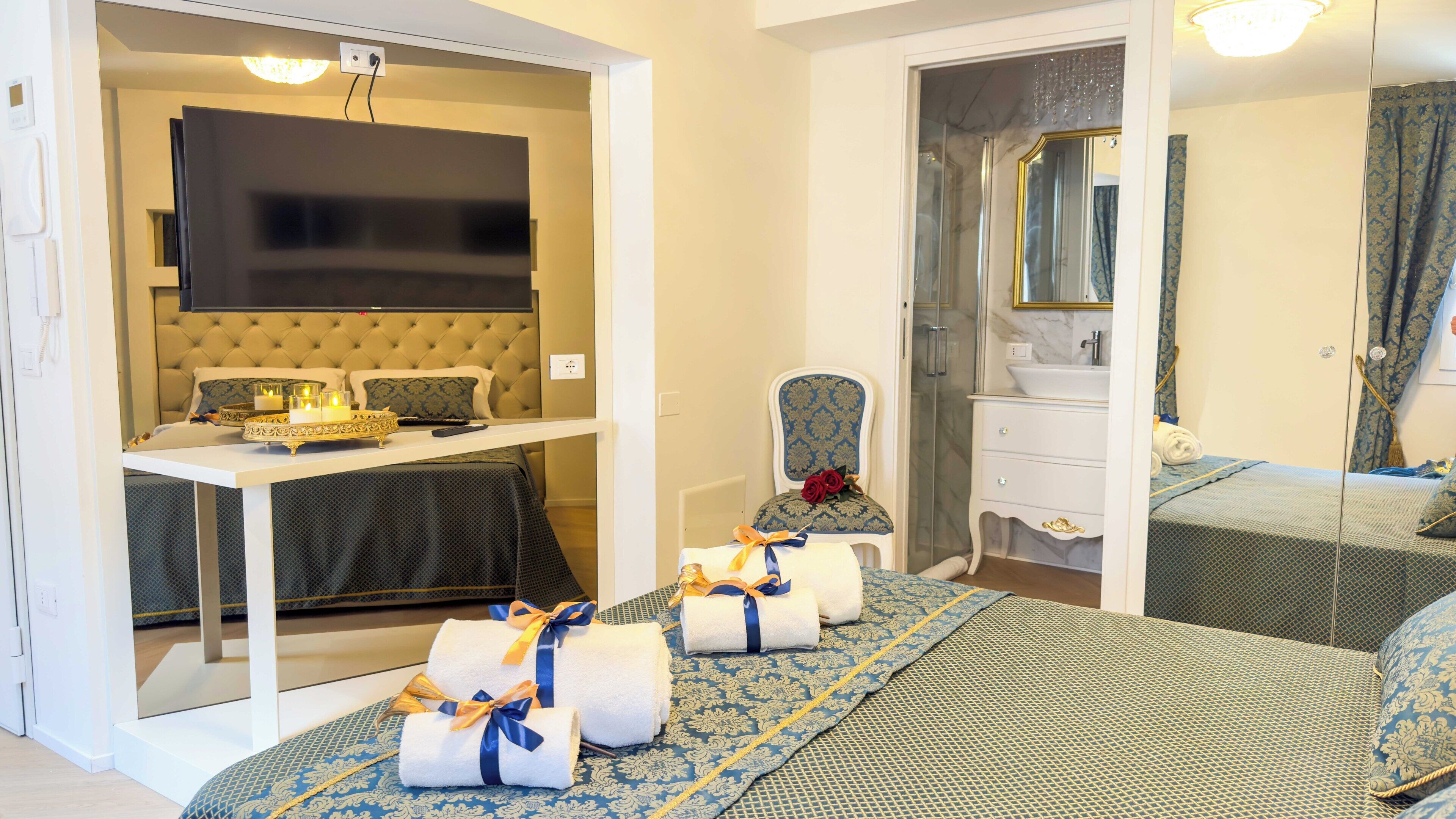 Ein Hotelzimmer mit Bett, einer Sitzecke mit Stuhl und Tisch, einem Badezimmer mit Spiegel und Waschbecken sowie einem an der Wand befestigten Fernseher.