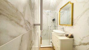 Un bany amb paret de marbre, un lavabo blanc, un mirall amb marc daurat i un espai de dutxa.