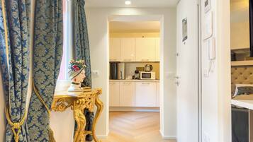 A room with a bed, a bedside table with a decorative item, a window with curtains, and a view into a kitchen area.