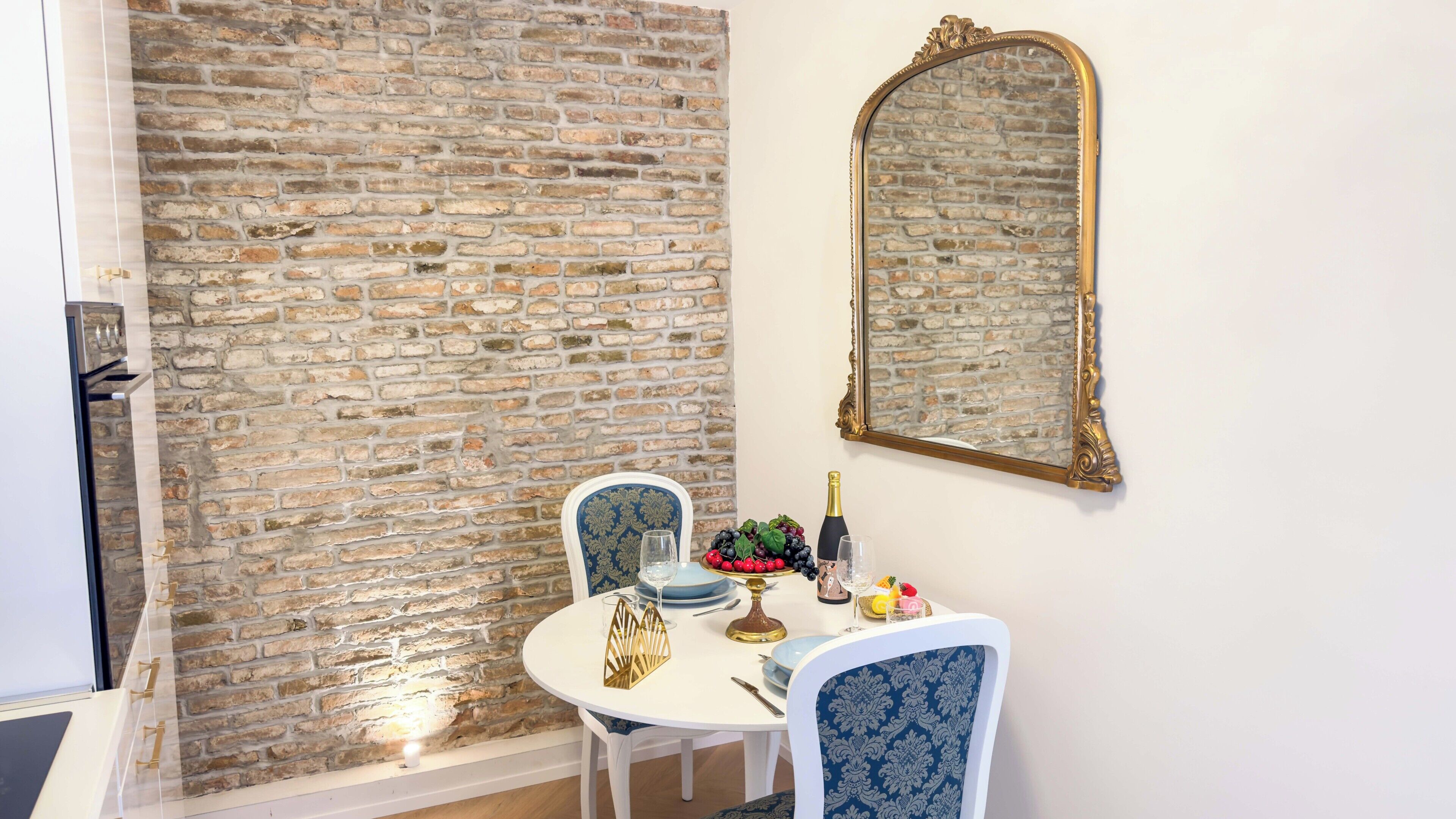 Un coin repas avec un mur en briques, un miroir et une table dressée avec des assiettes, des verres et un saladier de fruits.