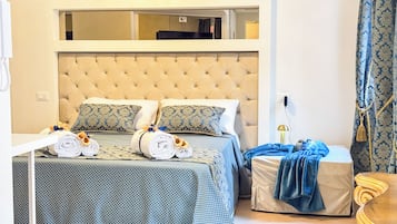 Une chambre d’hôtel avec une tête de lit capitonnée, deux lits avec une literie à motifs, une table de chevet avec des serviettes et une petite table de nuit avec une lampe.
