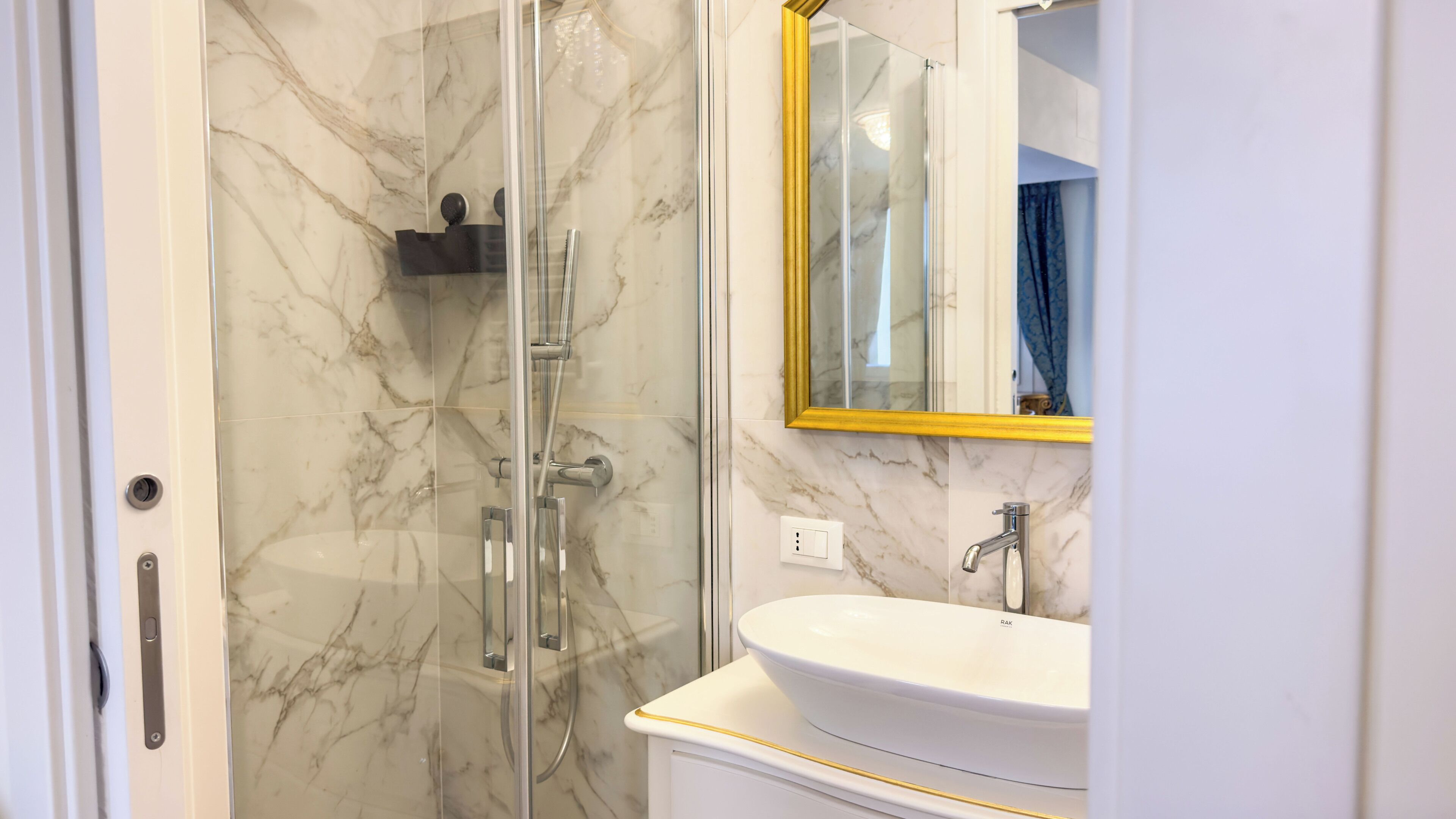 Un bagno con doccia vetrata, un lavandino bianco e uno specchio con cornice dorata.