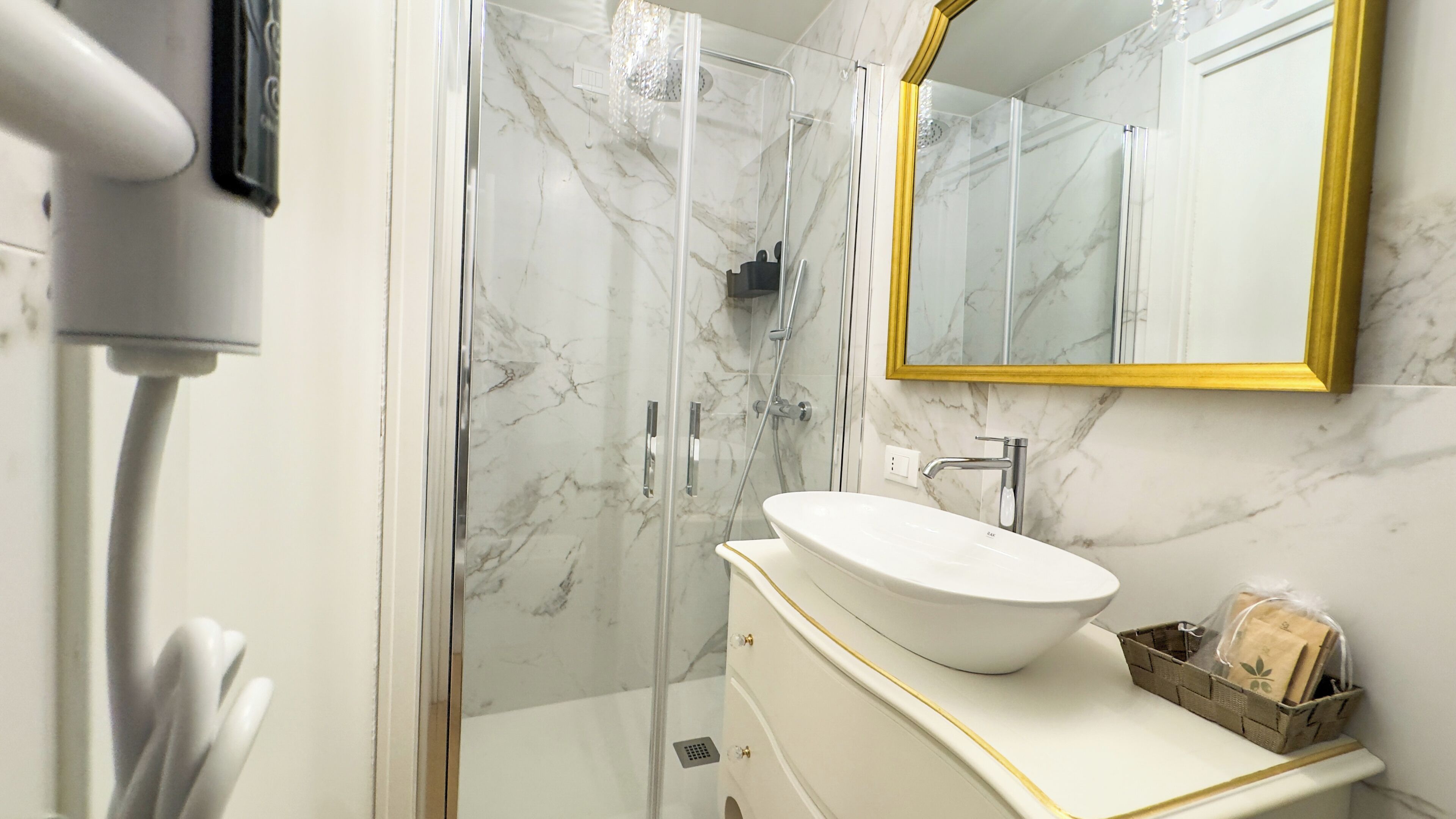 Un baño con lavamanos blanco, paredes de mármol, un espejo con marco dorado y cabina de ducha.
