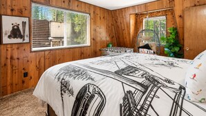 Een slaapkamer met houten wanden, een bed met een ski-thema dekbed, een raam met zonwering, een stoel en een plant.