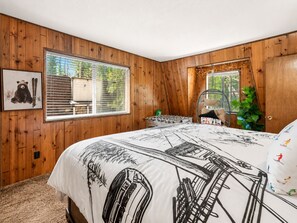 Ein Schlafzimmer mit Holzwänden, einem Bett mit Ski-Motiv auf der Tagesdecke, einem Fenster mit Jalousien, einem Stuhl und einer Pflanze.
