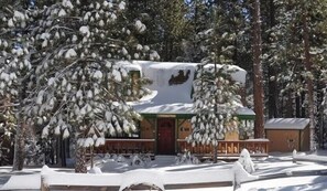 Una cabaña cubierta de nieve con techo verde, rodeada de árboles.
