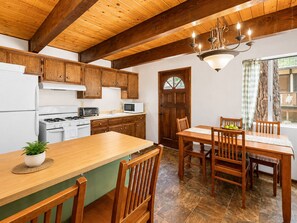 A kitchen with wooden cabinets, a stove, a microwave, and a dining area with a table and chairs. - Seasonal Creek Lodge - Cozy Forest Cabin w/ Hot Tub + Seasonal Creek (Big Bear City)
