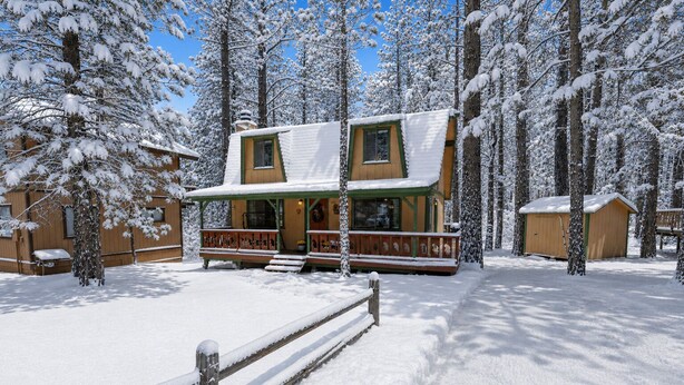 Een houten chalet met twee verdiepingen en een veranda, omringd door met sneeuw bedekte bomen.