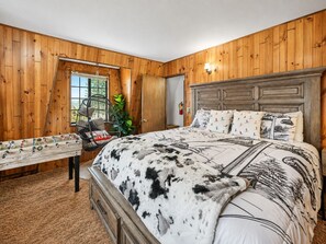 A bedroom with a wooden paneled wall, a bed with a patterned blanket, a hanging chair, and a window with a view. - Seasonal Creek Lodge - Cozy Forest Cabin w/ Hot Tub + Seasonal Creek (Big Bear City)