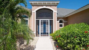 A building with a central entrance, a covered porch, and a well-maintained garden with palm trees and flowering shrubs. - Fish & Sips - Walking Distance to Cape Harbor - Kayak and Bikes - Intersecting Canal View (Cape Coral)