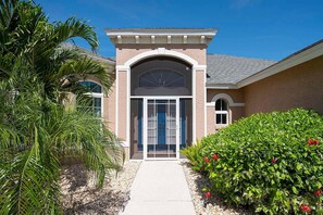 A building with a central entrance, a covered porch, and a well-maintained garden with palm trees and flowering shrubs. - Fish & Sips - Walking Distance to Cape Harbor - Kayak and Bikes - Intersecting Canal View (Cape Coral)