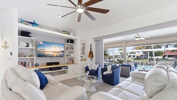 A spacious living room with a large TV, bookshelves, a ceiling fan, and a view of the outdoors. - Fish & Sips - Walking Distance to Cape Harbor - Kayak and Bikes - Intersecting Canal View (Cape Coral)