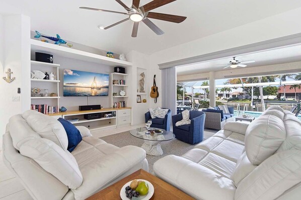 A spacious living room with a large TV, bookshelves, a ceiling fan, and a view of the outdoors. - Fish & Sips - Walking Distance to Cape Harbor - Kayak and Bikes - Intersecting Canal View (Cape Coral)