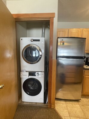 A compact laundry room with a stacked washer and dryer, a stainless steel refrigerator, and wooden cabinets. - 2-bedroom resort in welcoming Breckenridge. Nov 30th-Dec 5th, 2026. (Breckenridge)