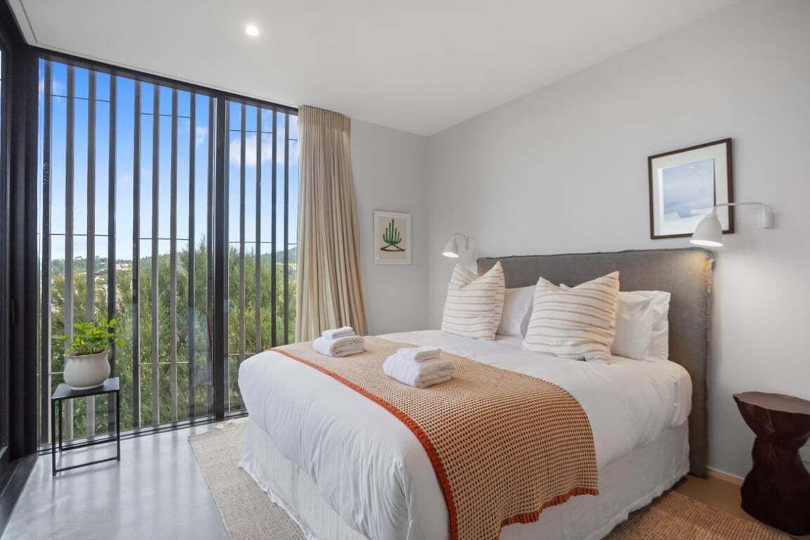 A modern bedroom with a large bed, a view through a sliding glass door, a bedside table with a plant, and a framed picture on the wall.